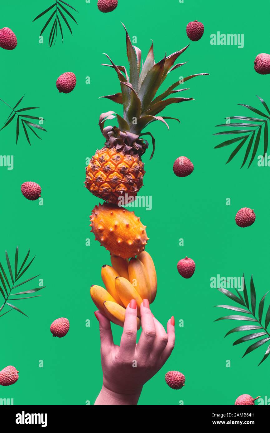 Surtido de frutas tropicales, pirámide balanceando o mano humana sobre fondo verde. Piña, kiwano, kiwi, lichee y plátano - torre de exótico Foto de stock