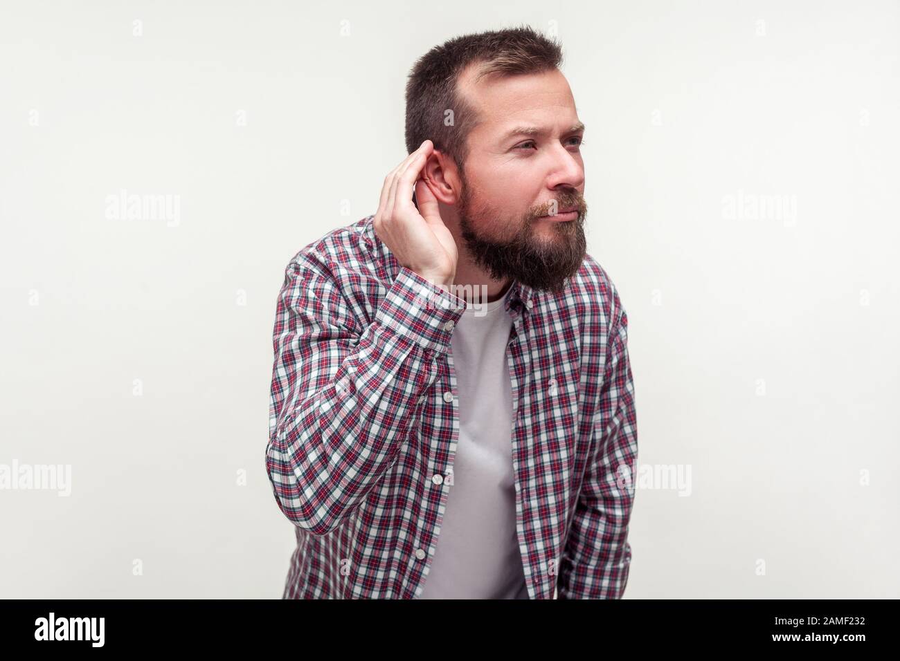 Retrato del hombre sordo con barba en plaid camisa de pie con la mano cerca del oído y escuchando cuidadosamente la conversación secreta, problemas de audición, sordera. In Foto de stock