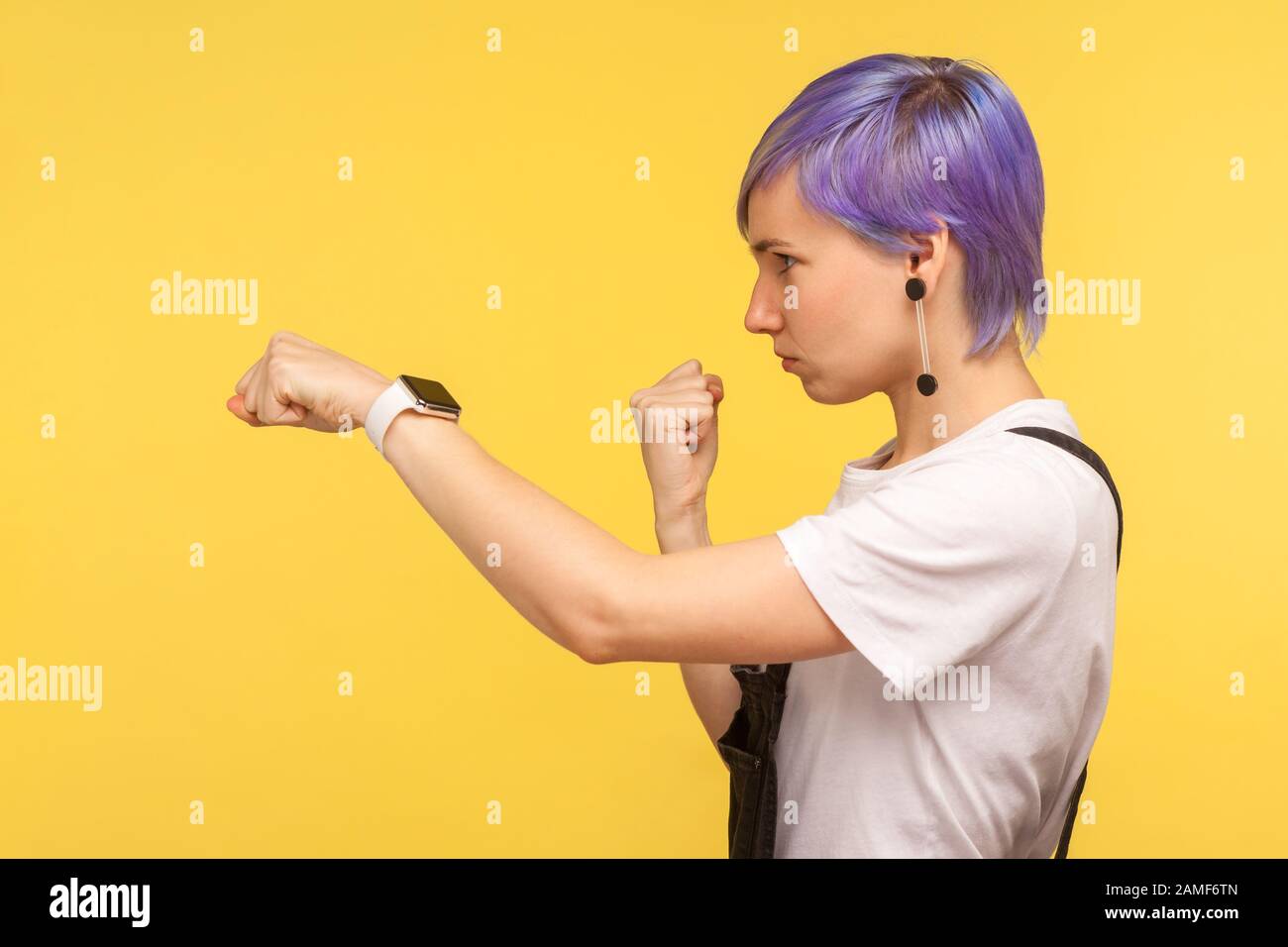 Vista Lateral De Una Determinada Mujer Joven Hipster Con Pelo Corto Tenido Violeta En Monos Denim Golpeando Con Puno Mostrando El Gesto De Boxeo Mirando A La Confitura Fotografia De Stock