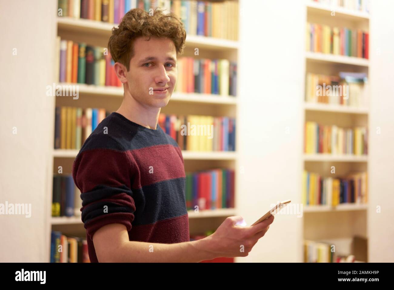 Adolescente en la biblioteca permaneciendo delante de la estantería con el smartphone en su mano Foto de stock
