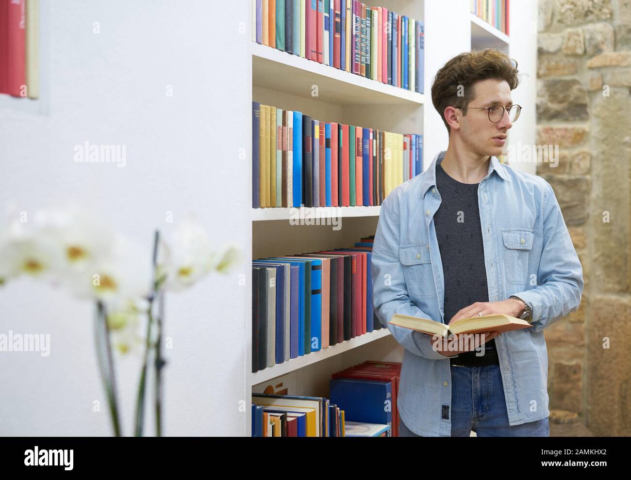Estudiante joven con gafas en la biblioteca mirando a su lado izquierdo y sosteniendo un libro en sus manos. Foto de stock
