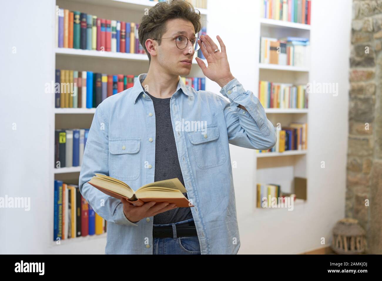 Estudiante joven con libro en su mano de pie delante de una estantería comprobando sus gafas y mirando hacia arriba. Foto de stock