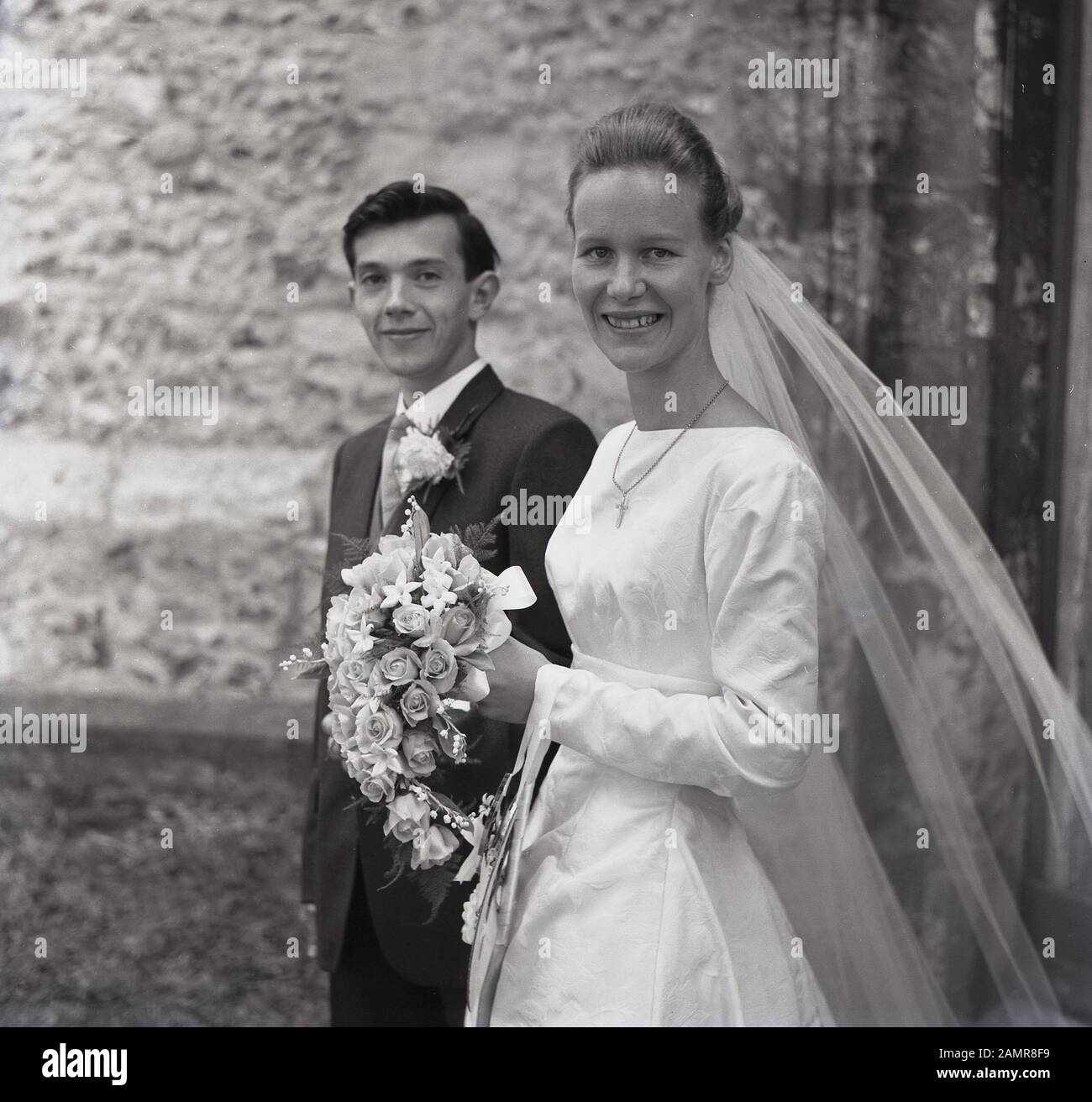 1960, pareja de bodas de adultos jóvenes, novia sonriente en su vestido de boda, con velo sosteniendo un ramo de flores fuera de una iglesia, Inglaterra, Reino Unido. Foto de stock