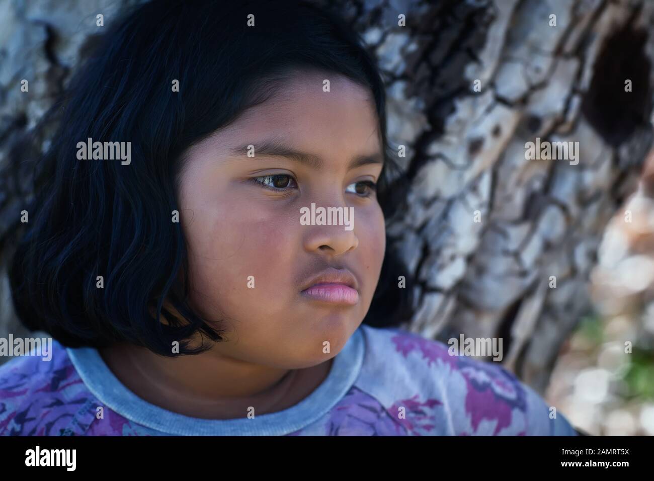 Una niña con una expresión facial molesta de pie delante de un árbol. Foto de stock
