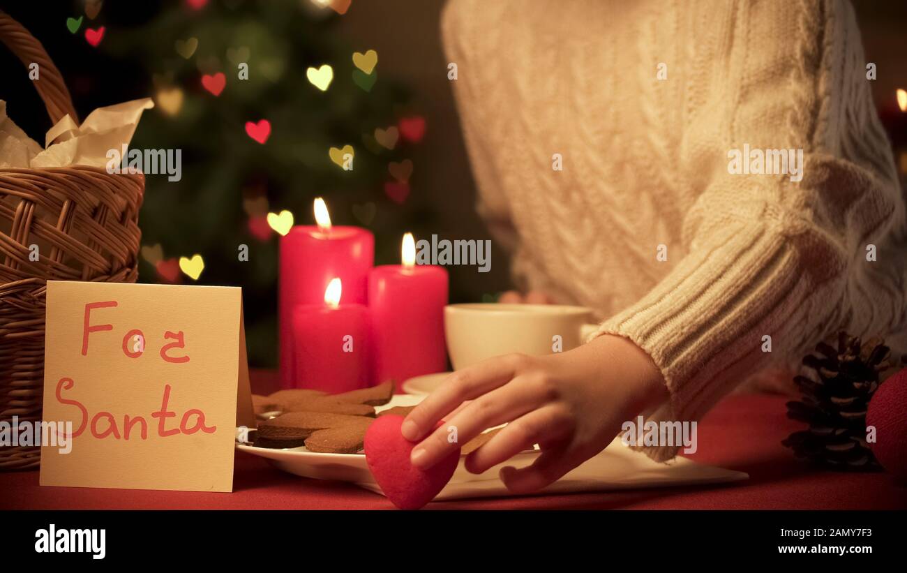 Chica colocando corazón de juguete cerca de la decoración de Navidad, chocolate caliente y sabrosa galleta de jengibre Foto de stock