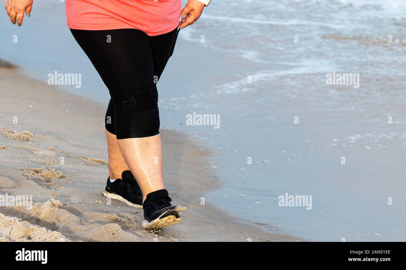Una gran mujer de la cintura para abajo está caminando en la playa, junto al agua para hacer ejercicio vistiendo spandex y zapatos para correr. Foto de stock