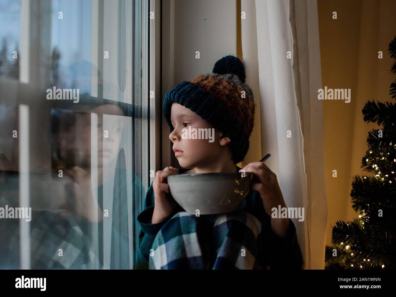 el niño se sentó en un alféizar de la ventana con un cuenco de cereales mirando hacia fuera Foto de stock