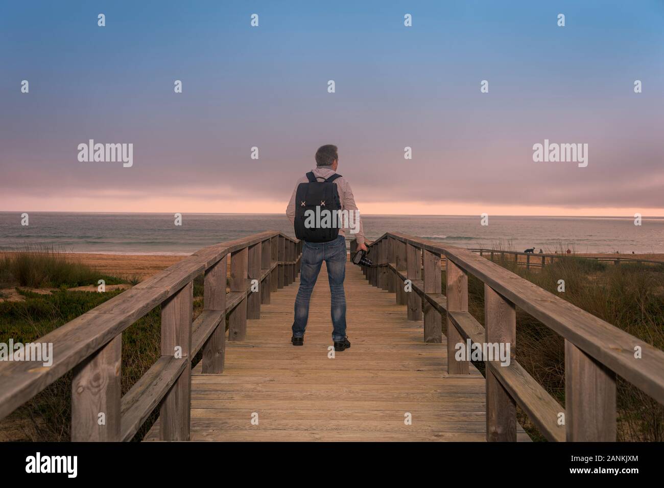 Vista posterior de un fotógrafo con una mochila de pie sobre una pasarela de madera con vistas al océano. Foto de stock