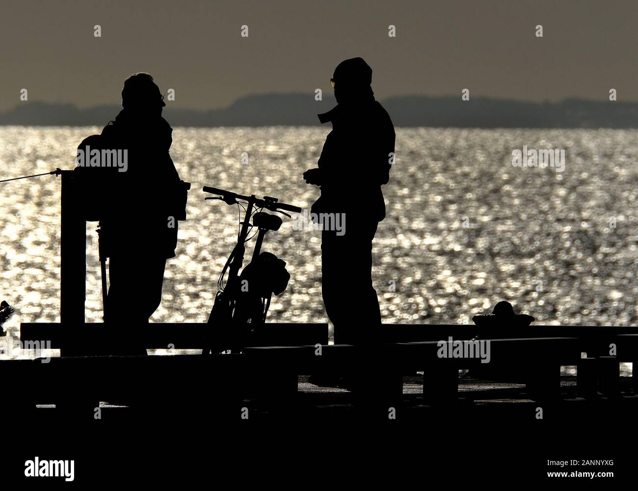 Las personas en silueta y el sol brillante en el extremo de un muelle junto al mar. Foto de stock
