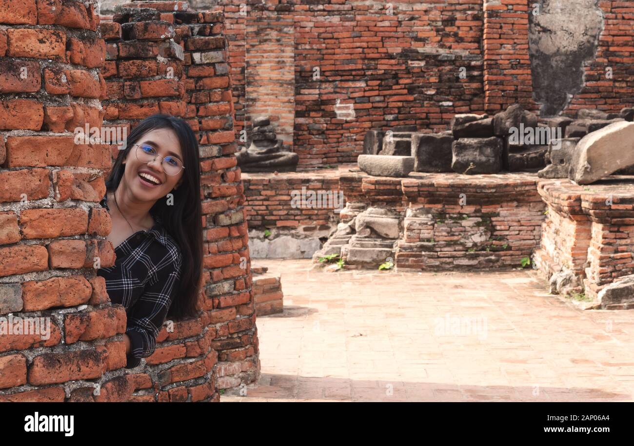 Diversión amante joven diversa niña asiática escondiéndose detrás de pared de ladrillo y sonriendo - Milennial hipster social influencer viajar a destinos turísticos culturales - viaje, tendencias y ideas de vacaciones concepto Foto de stock