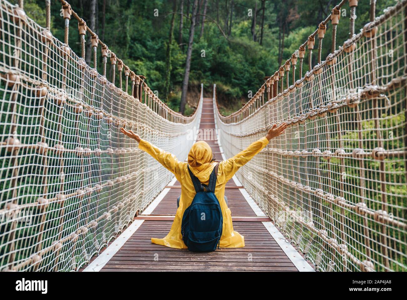 Mujer excursionista con mochila y impermeable amarillo sentado en el puente colgante. Foto de stock