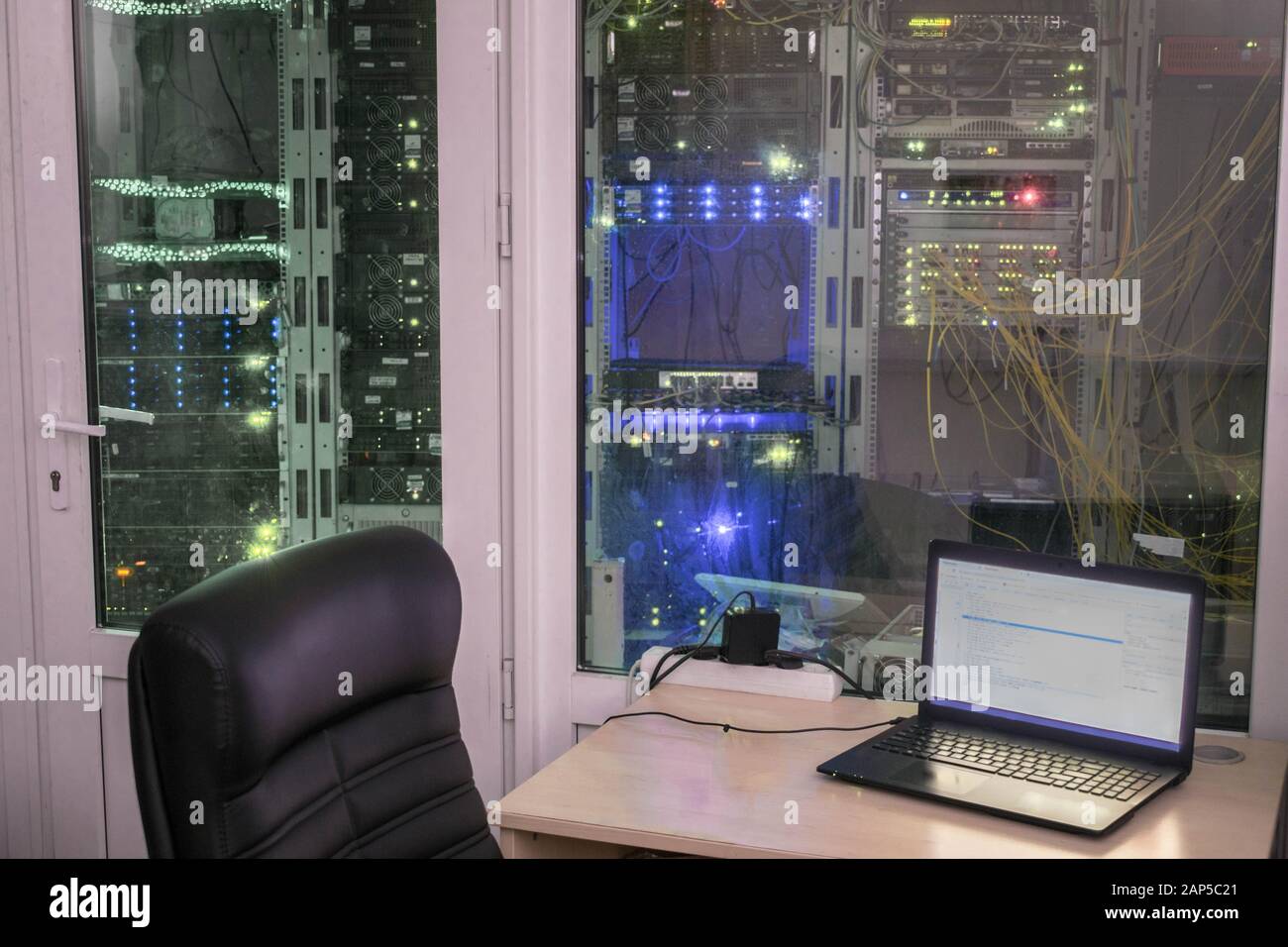 Muchos racks con equipos informáticos están detrás de la pared de cristal de un moderno centro de datos. El portátil está sobre la mesa frente a potentes routers. Vaciar trabajo Foto de stock