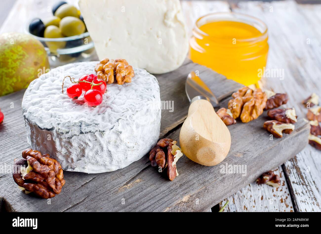 Diferentes tipos de quesos deliciosos en una tabla de madera y muchos aperitivos, nueces, miel, bayas, aceitunas en una mesa de madera blanca vieja. Ingredientes de la comida de vuelta Foto de stock