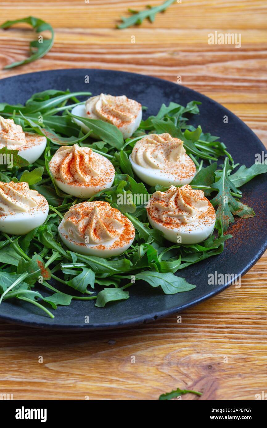 Huevos devanados con pimentón en ensalada de rúcula fresca en plato rural negro sobre mesa de madera, aperitivo vegetariano saludable o aperitivo de cerca Foto de stock