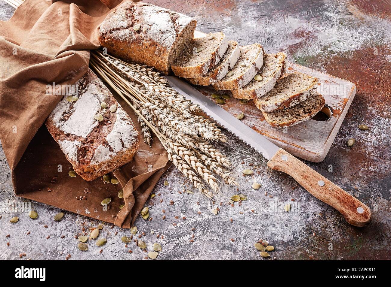 Pan recién horneado con semillas de girasol y calabaza en una servilleta marrón. Pan en rebanadas en una tabla de madera y un cuchillo de cocina. Orejas de trigo. Foto de stock