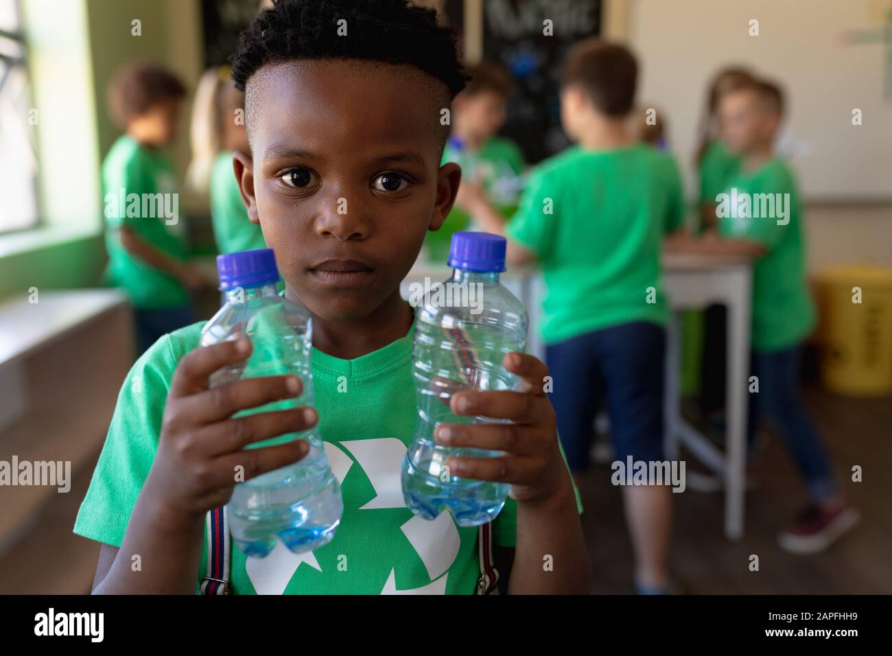 Niño escolar que lleva una camiseta verde con un logotipo de reciclaje blanco en ella y que sostiene dos aguas de plástico bo Foto de stock