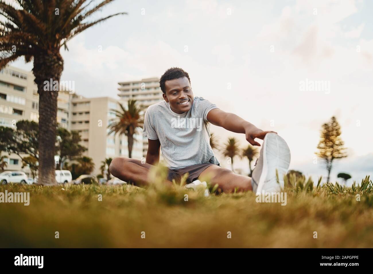 Retrato sonriente de un joven atleta sentado en hierba verde haciendo ejercicio de estiramiento en el parque Foto de stock