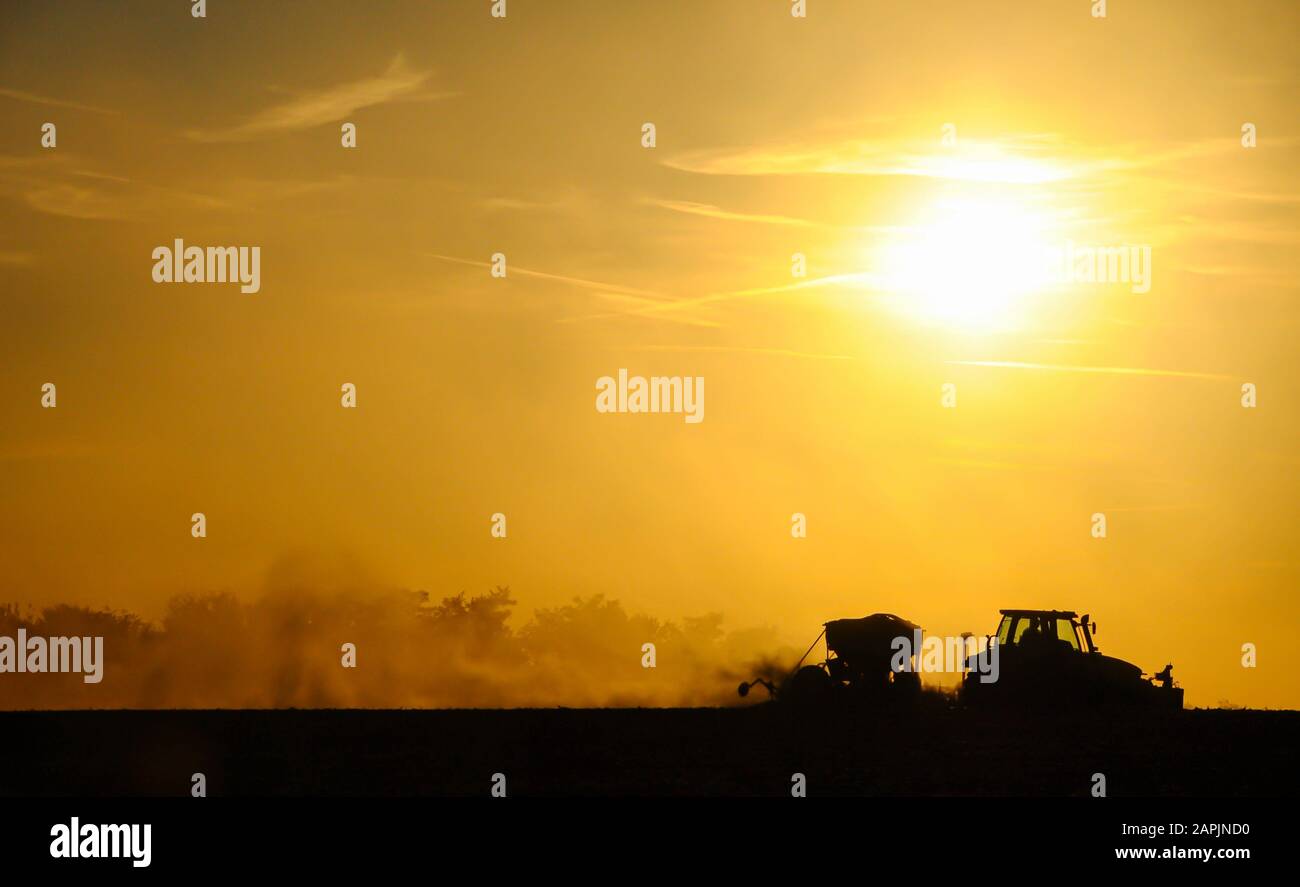 Silueta de un tractor sembrando semillas en un campo en una nube de polvo contra el fondo de la puesta de sol. Foto de stock