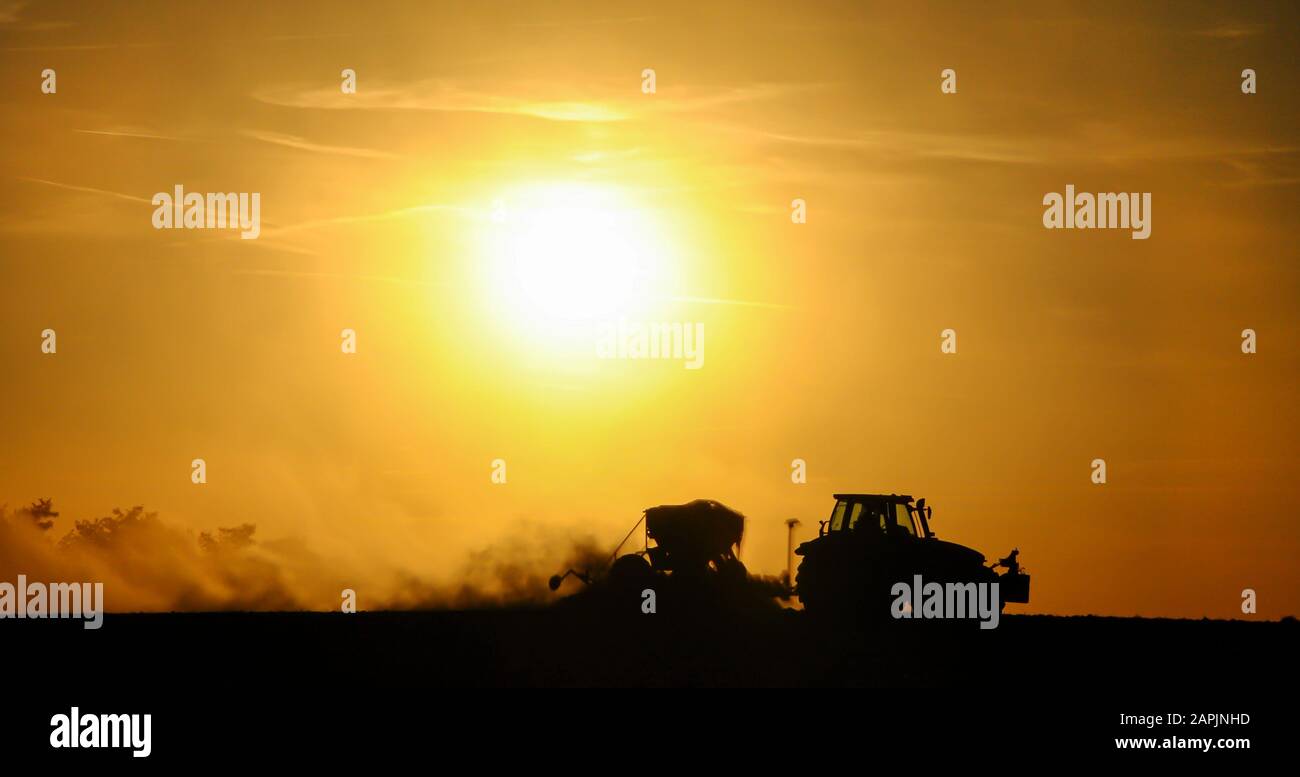 Silueta de un tractor sembrando semillas en un campo en una nube de polvo contra el fondo de la puesta de sol. Foto de stock