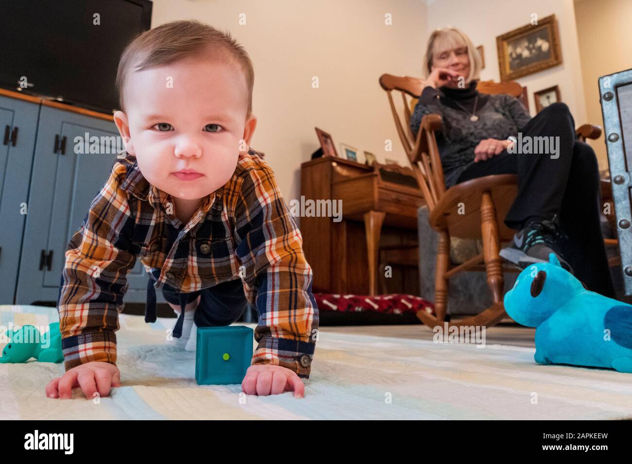 Niño de siete meses felizmente arrastrándose en la sala de estar; abuela sentada en el fondo Foto de stock