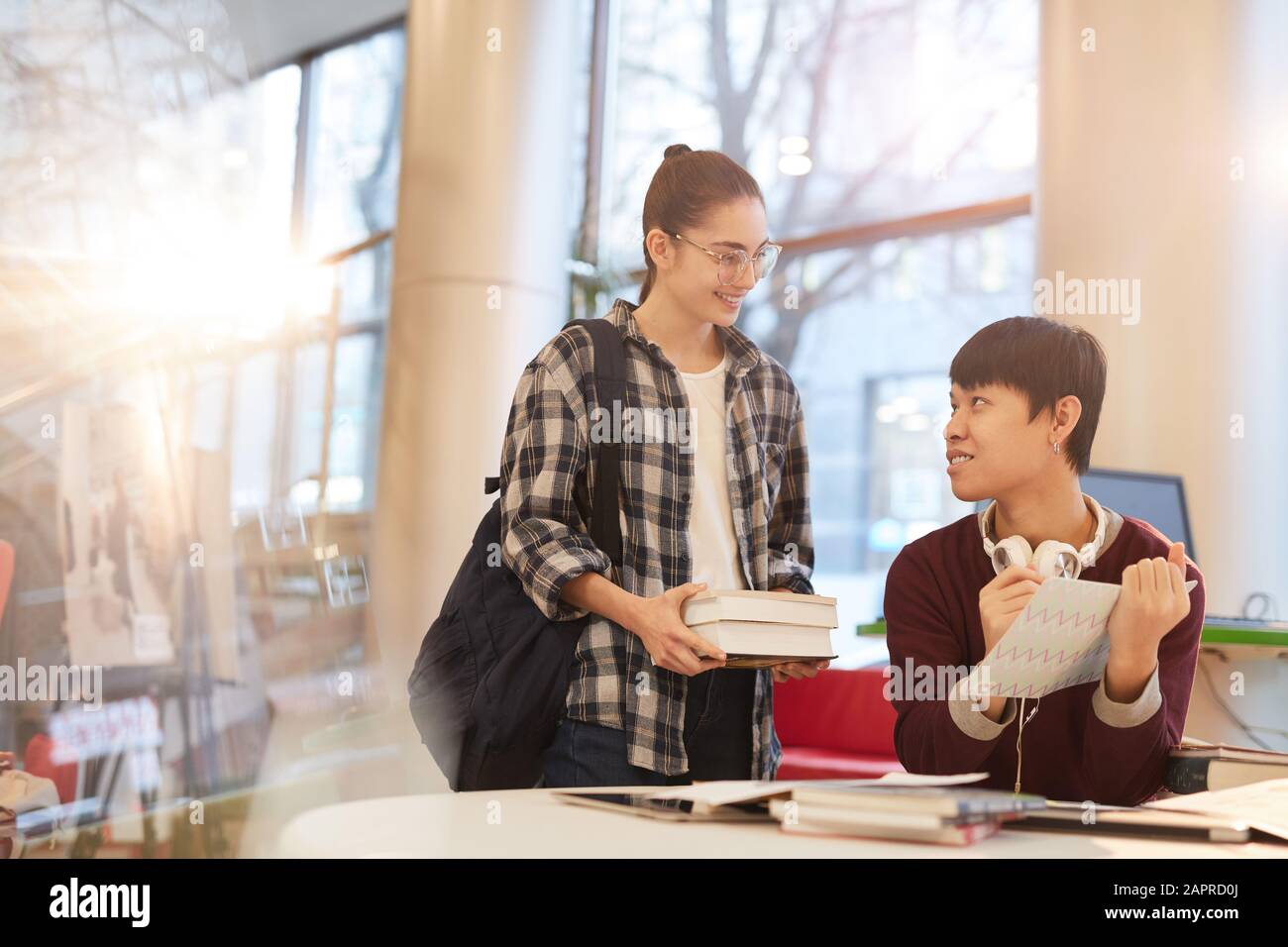 Joven adolescente sonriente sosteniendo libros y hablando con su compañero de clase que estudian en la biblioteca juntos Foto de stock