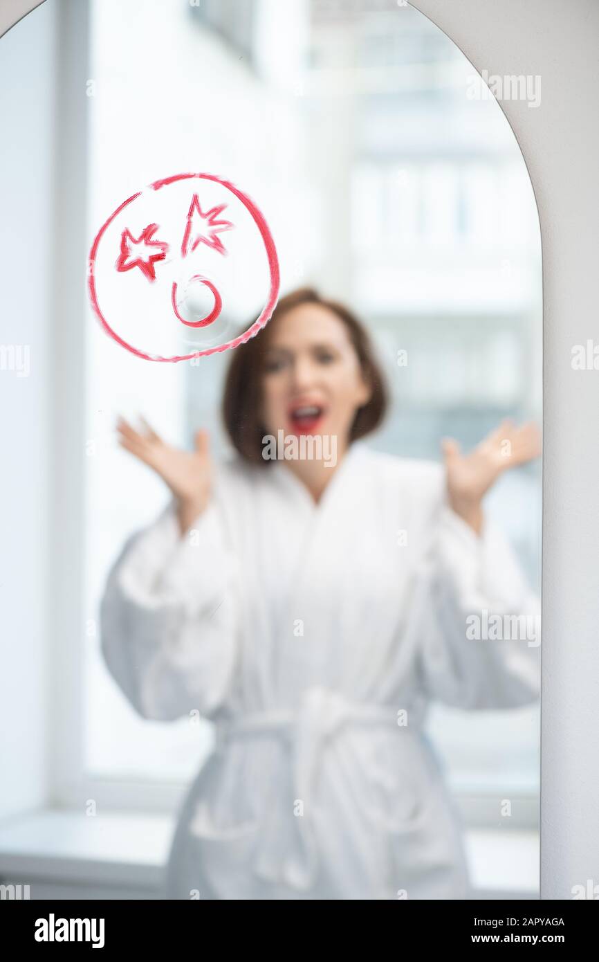 Una joven mujer gritando frente al espejo con una cara conmocionada dibujada con lápiz labial Foto de stock