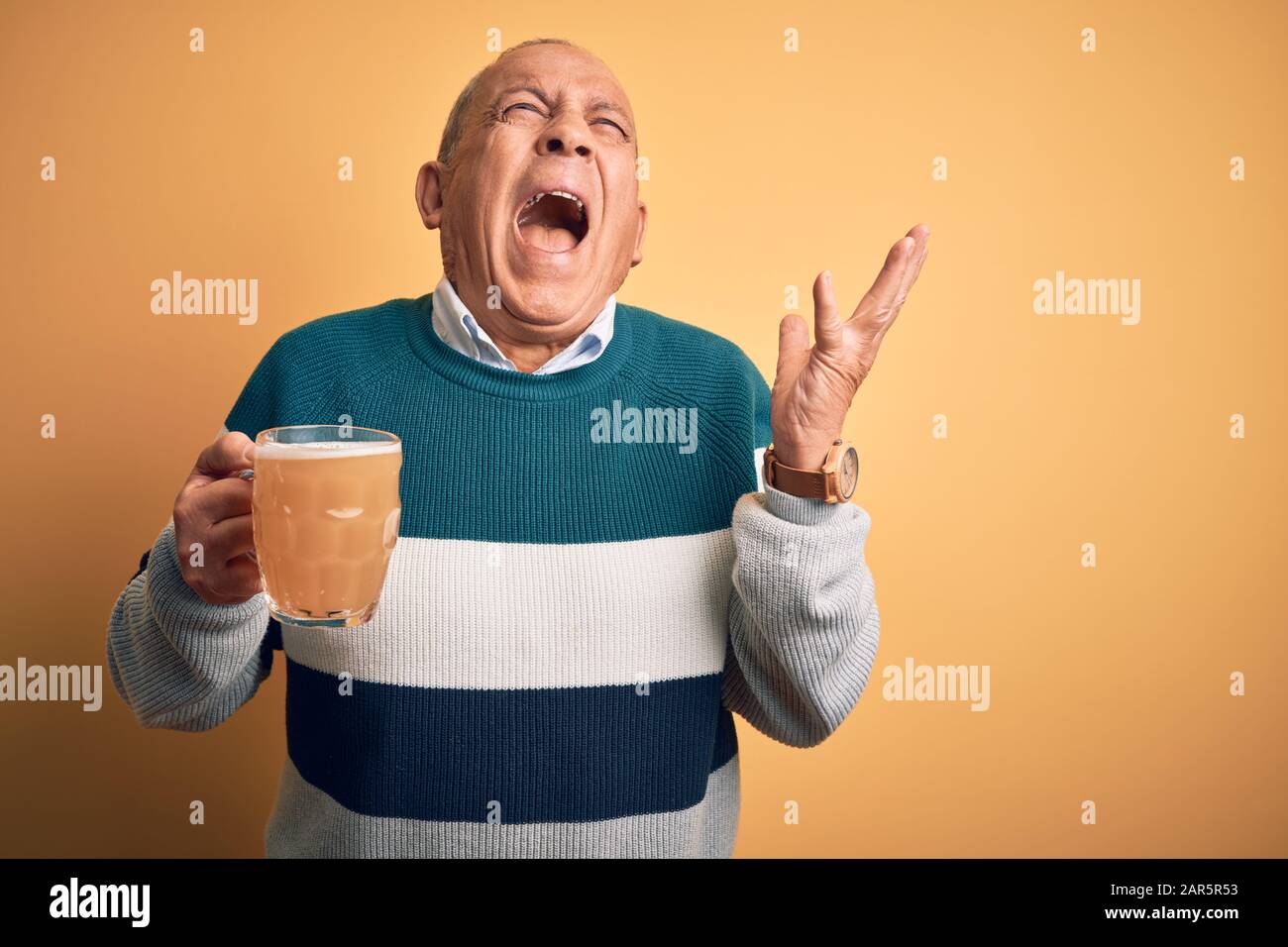 Hombre guapo mayor bebiendo tarro de cerveza de pie sobre fondo amarillo aislado loco y loco gritando y gritando con expresión agresiva y ar Foto de stock