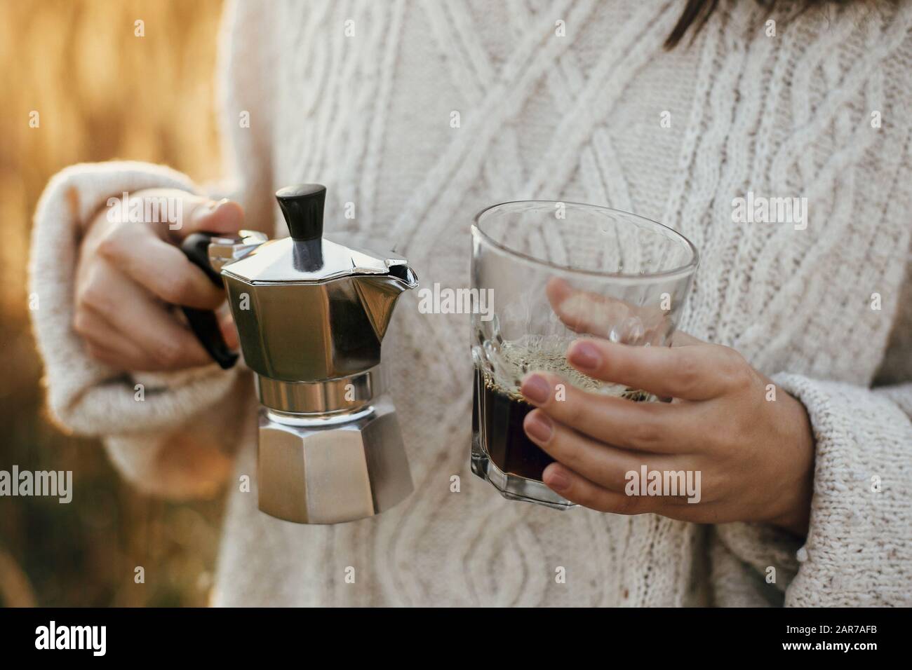 Hipster mujer que sostiene géiser cafetera y taza de cristal con café en la  luz cálida y soleada en las hierbas rurales del campo. Momento rústico  atmosférico. Alterno Fotografía de stock -