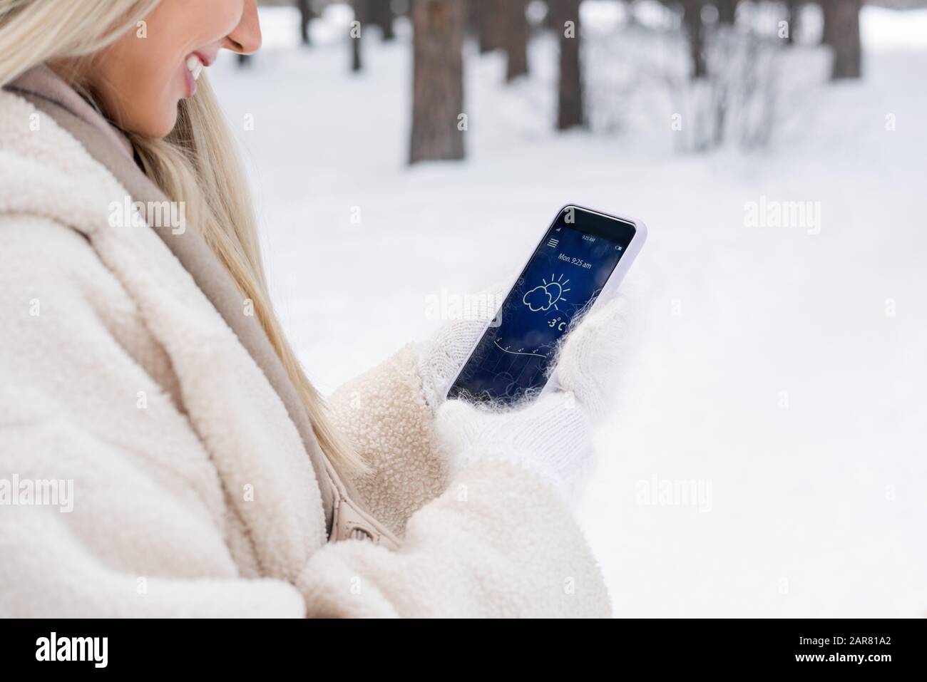 Mujer joven rubia con abrigo blanco de invierno y manoplas de punto con smartphone Foto de stock