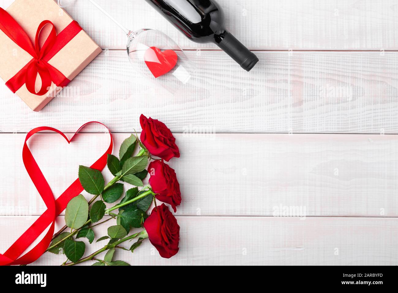 Día de San Valentín cena romántica, cinta roja, kraft caja de regalo,  corazón en copa de vino, botella, rosas, sobre fondo blanco de madera.  Espacio de copia, lugar fo Fotografía de stock -