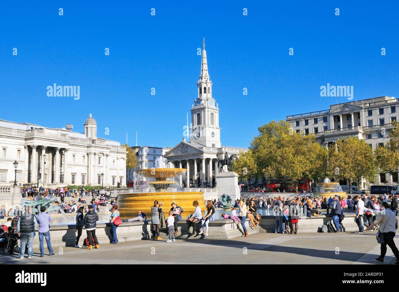 Turistas sentados alrededor de las fuentes en una soleada Plaza Trafalgar con San Martín en La iglesia De Los Campos en el fondo, Londres, Inglaterra, Reino Unido Foto de stock