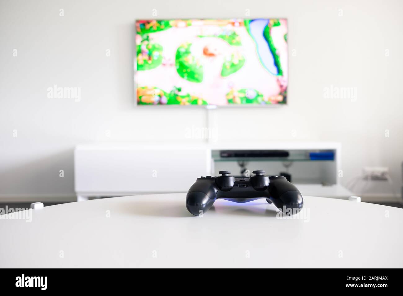 Vista de un controlador de videojuegos negro sin logotipo en una mesa blanca. El televisor está en segundo plano. Foto de stock