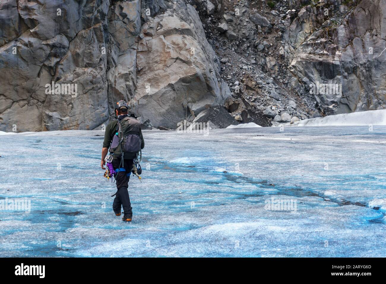 El escalador profesional de hielo está caminando por el glaciar. Senderismo en el hielo. Montaña de roca gris sobre un fondo. El hombre lleva todo el equipo y casco. Foto de stock