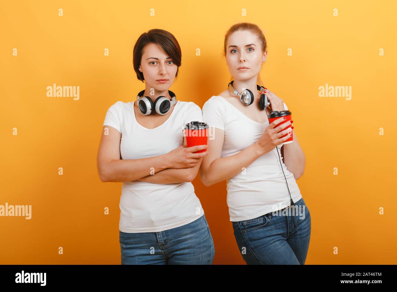 Dos mujeres serias con auriculares sosteniendo un cristal de café de papel y mirando la cámara - Imagen Foto de stock