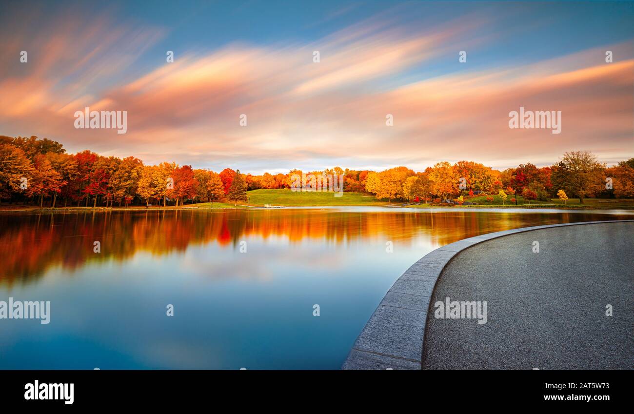 El hermoso follaje naranja del otoño y el cielo azul con nubes llenas de color y rayas se reflejan en la superficie perfectamente quieta del lago. Foto de stock