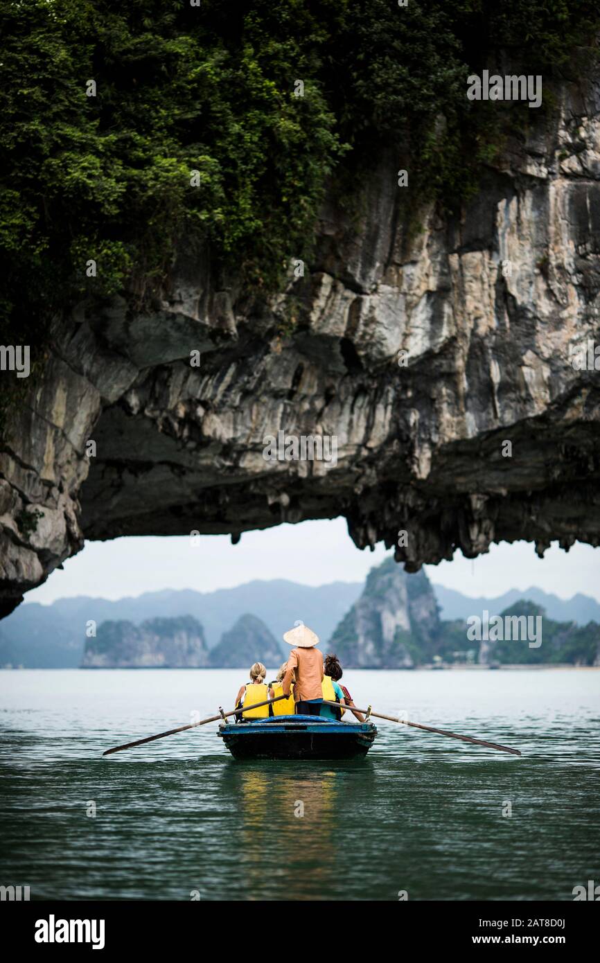 Vista trasera del hombre usando sombrero de paja que transporta a un pequeño grupo de personas en un barco, remando debajo del arco de roca natural. Foto de stock