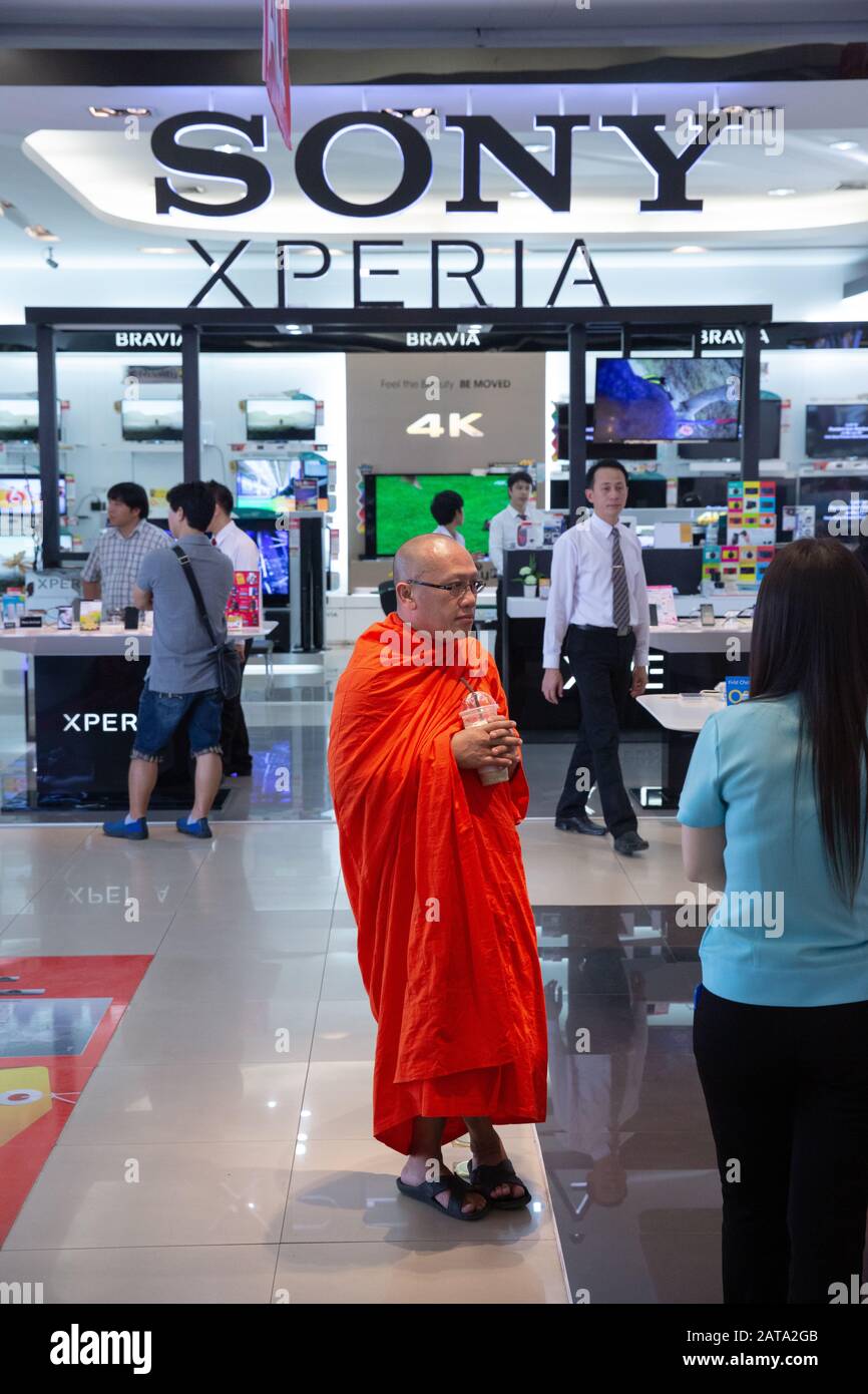 Monje budista en estría naranja y sandalias entre televisores 4K y otros aparatos electrónicos de alta tecnología en un centro comercial en Chiang Mai, Tailandia. Foto de stock