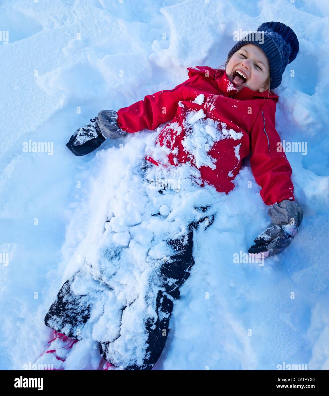 Niña jugando ángel de nieve, niño alegre tumbado en la nieve y gritando de alegría, diversión al aire libre, disfrutando de las vacaciones de invierno, feliz infancia Foto de stock