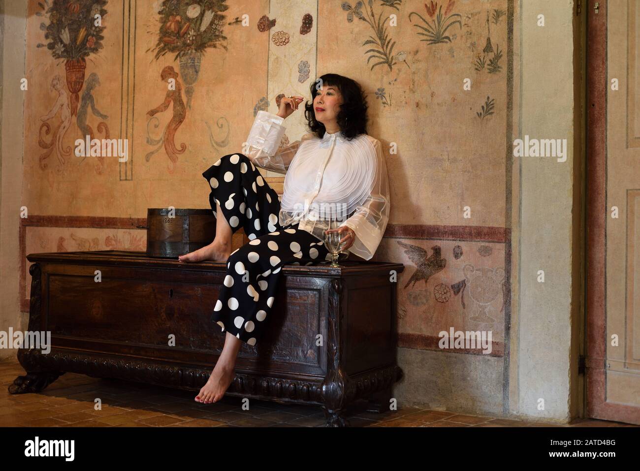 Una mujer sofisticada y elegante glamurosa sentada en un baúl antiguo en una lujosa sala de castillo medieval con trompe l'oeil pintado Foto de stock