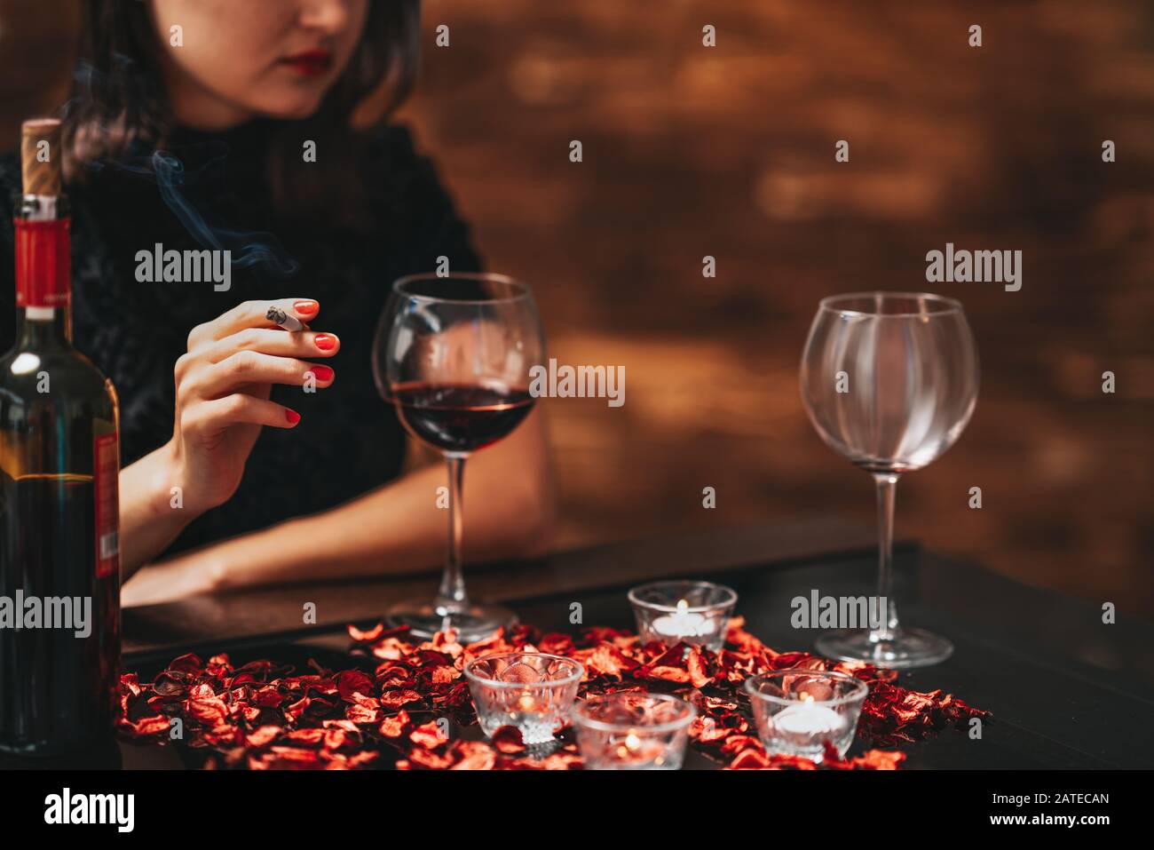 Sola y infeliz hermosa mujer sentado en una mesa, beber vino y fumar con un mal humor Foto de stock