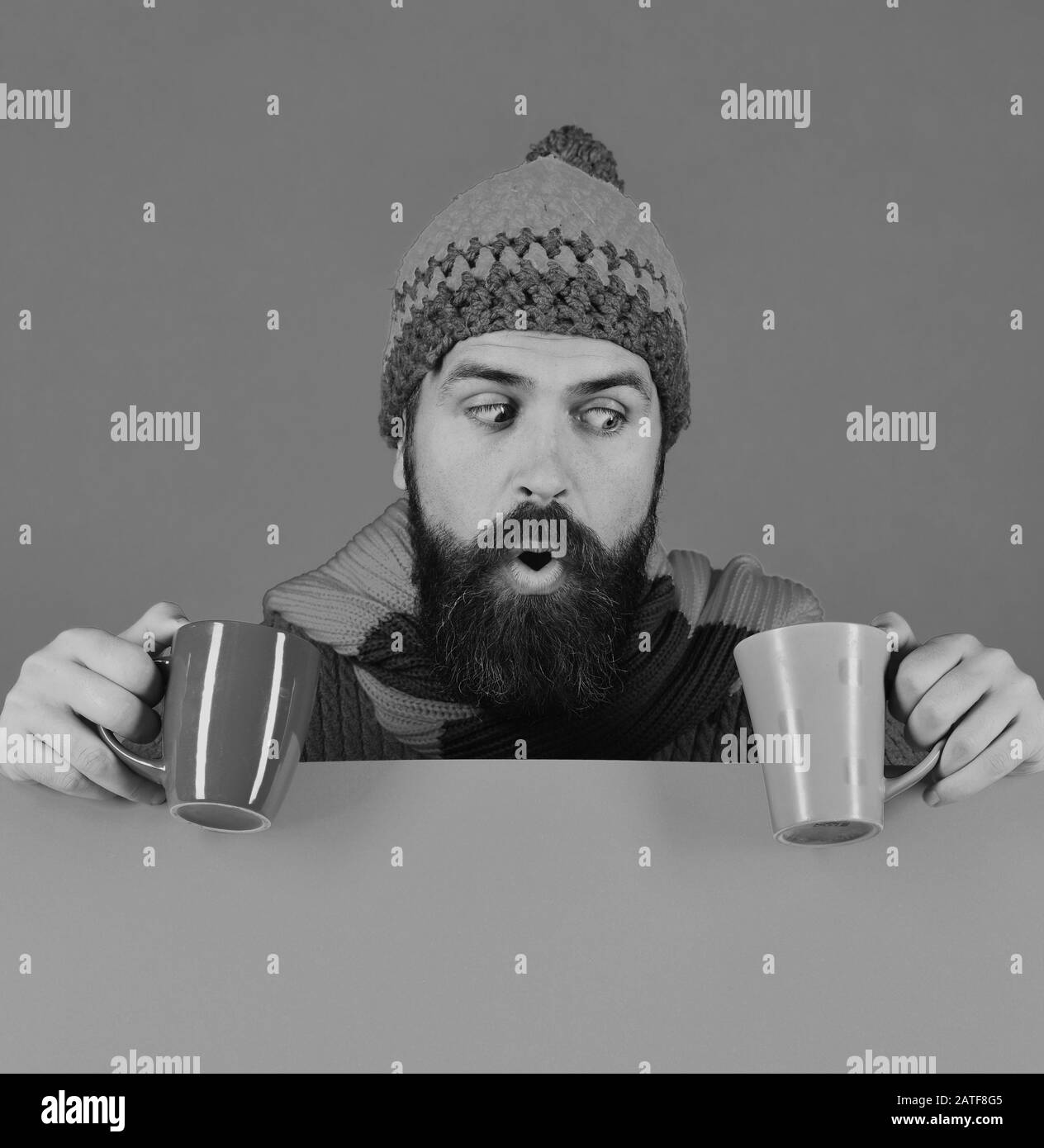 El hombre con sombrero cálido sostiene tazas marrones y verdes sobre fondo verde y naranja, espacio de copia. Hipster con barba y cara conmocionada tiene una taza de café. Idea de tiempo de desayuno. Perfecto concepto de aroma y mañana de otoño Foto de stock