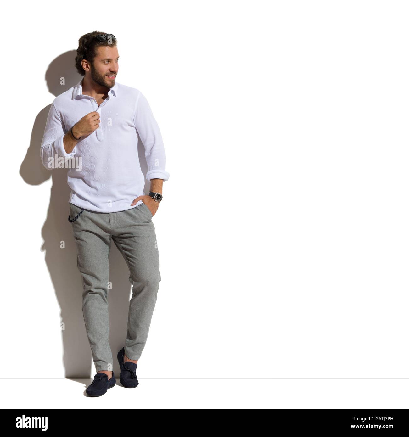 Un hombre guapo con camisa blanca, pantalones grises y mocasines está  parado contra la pared soleada con la mano en el bolsillo y mirando al  lado. Vista frontal. Completo Fotografía de stock -