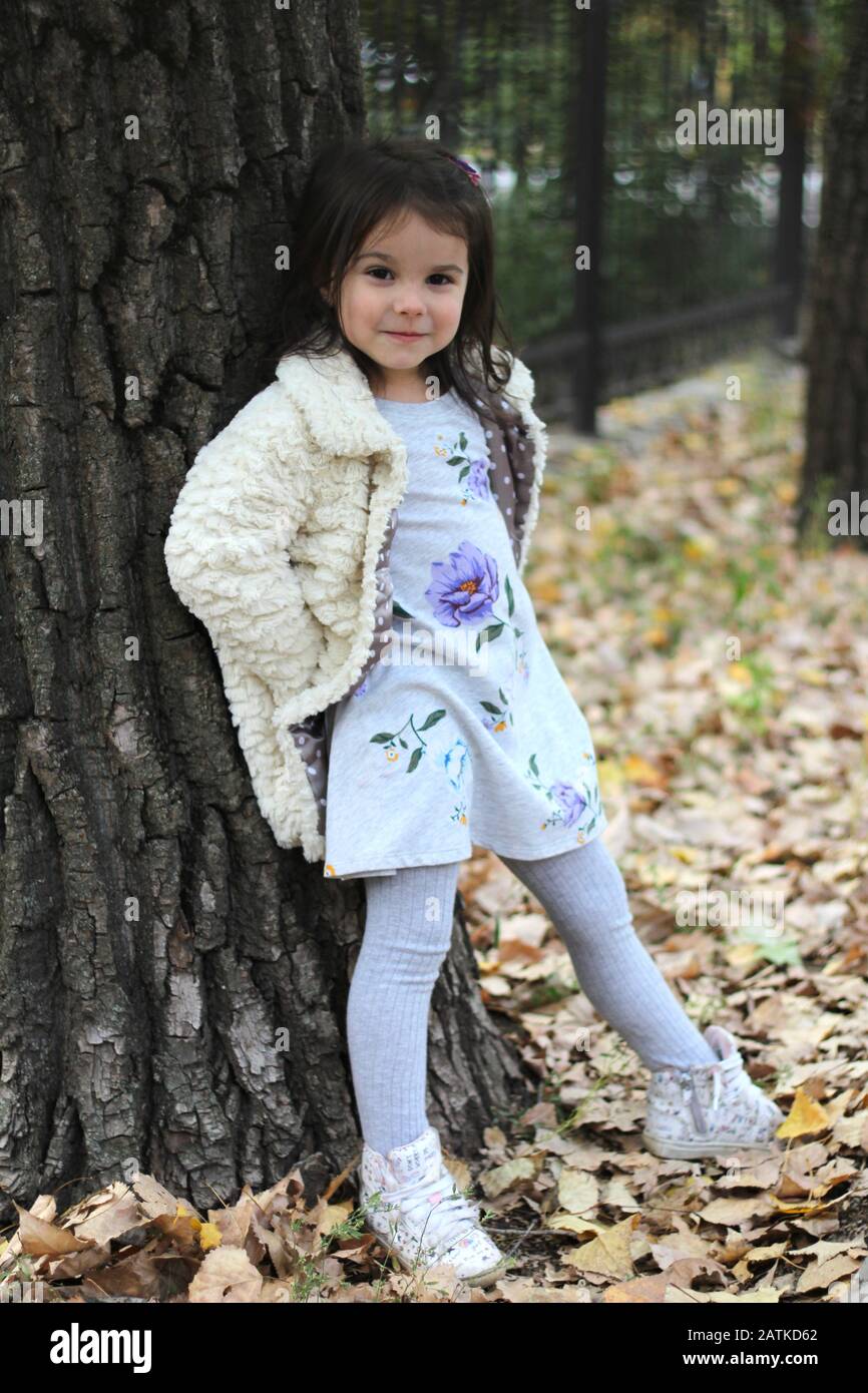 Una niña con pelo largo y oscuro un suave abrigo blanco y vestido colorido está de pie un gran árbol. Fondo de parque difuminado de stock - Alamy
