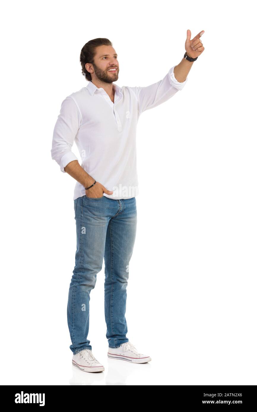 Un hombre guapo con camisa blanca, jeans sneakers está de pie con la mano en el mirando hacia arriba y apuntando. Vista frontal lateral. Estudio de longitud completa con ISO