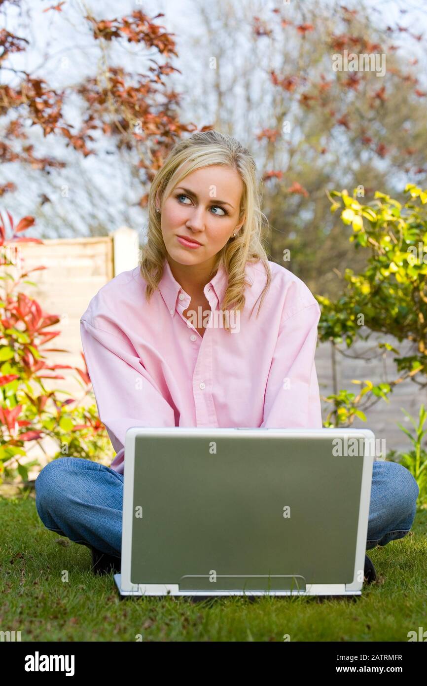 Hermosa mujer joven adolescente sonriente pensativa sentada en su jardín en casa utilizando un ordenador portátil para compras en línea o medios sociales Foto de stock