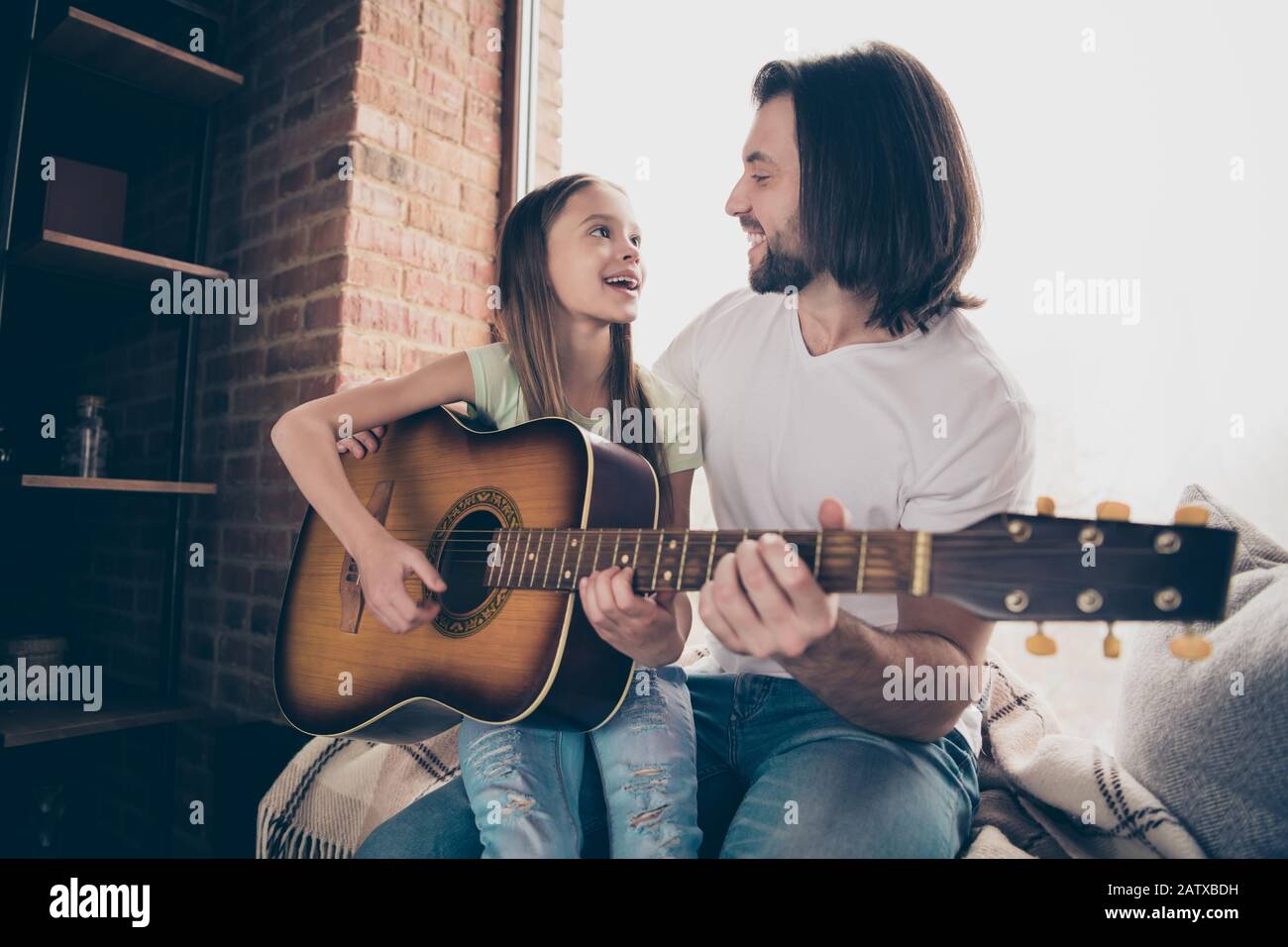 Foto de la pequeña adorable dama bonita y su guapo joven papá sentarse ventana bajo los ojos mantener la guitarra acústica aprender nueva canción pasar tiempo Foto de stock