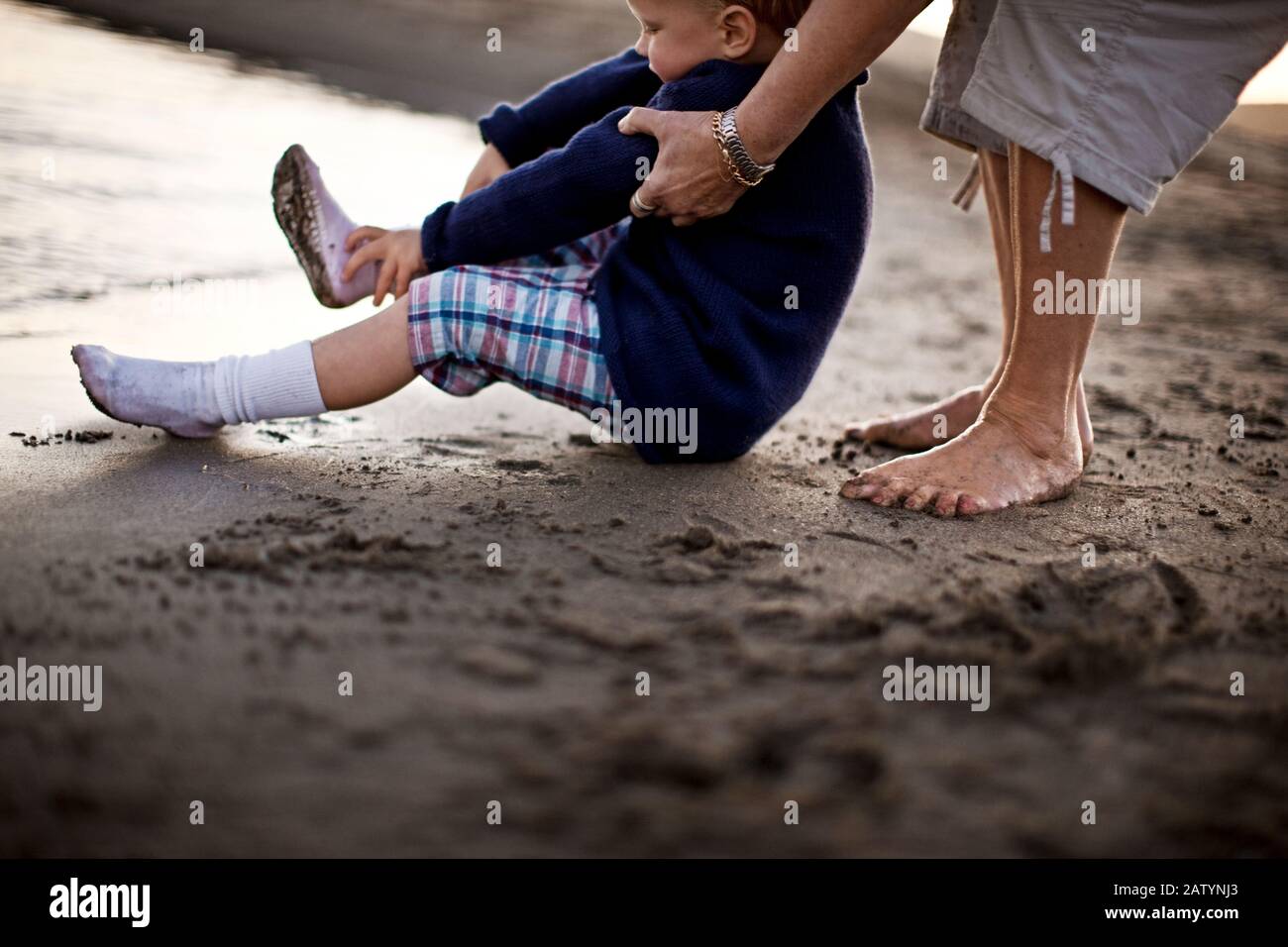 Mujer sosteniendo a un niño pequeño en la playa mientras se quita los calcetines para ir a Fotografía stock Alamy