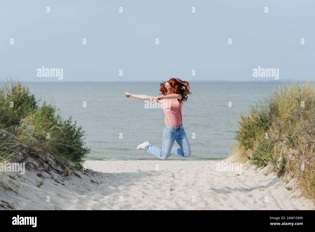 Mujer joven sin preocupaciones saltando para la alegría en un camino de arena a través de las dunas sobre una playa tropical Foto de stock