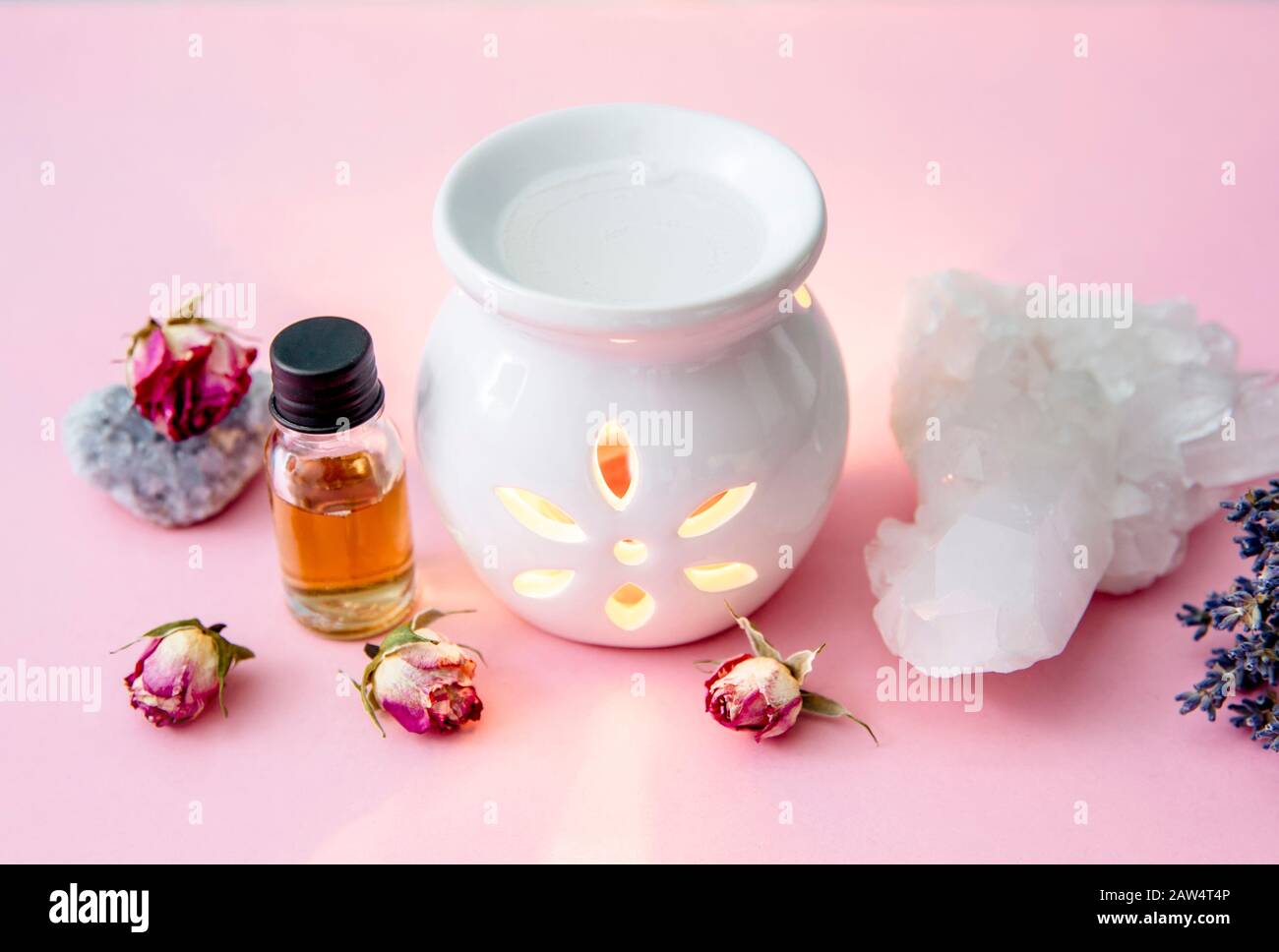 Lámpara de aceite de aroma de vela blanca cerámica con botella de aceite esencial y flores secas, geodes de cristal en un moderno fondo rosa pastel y azul en el interior. Foto de stock