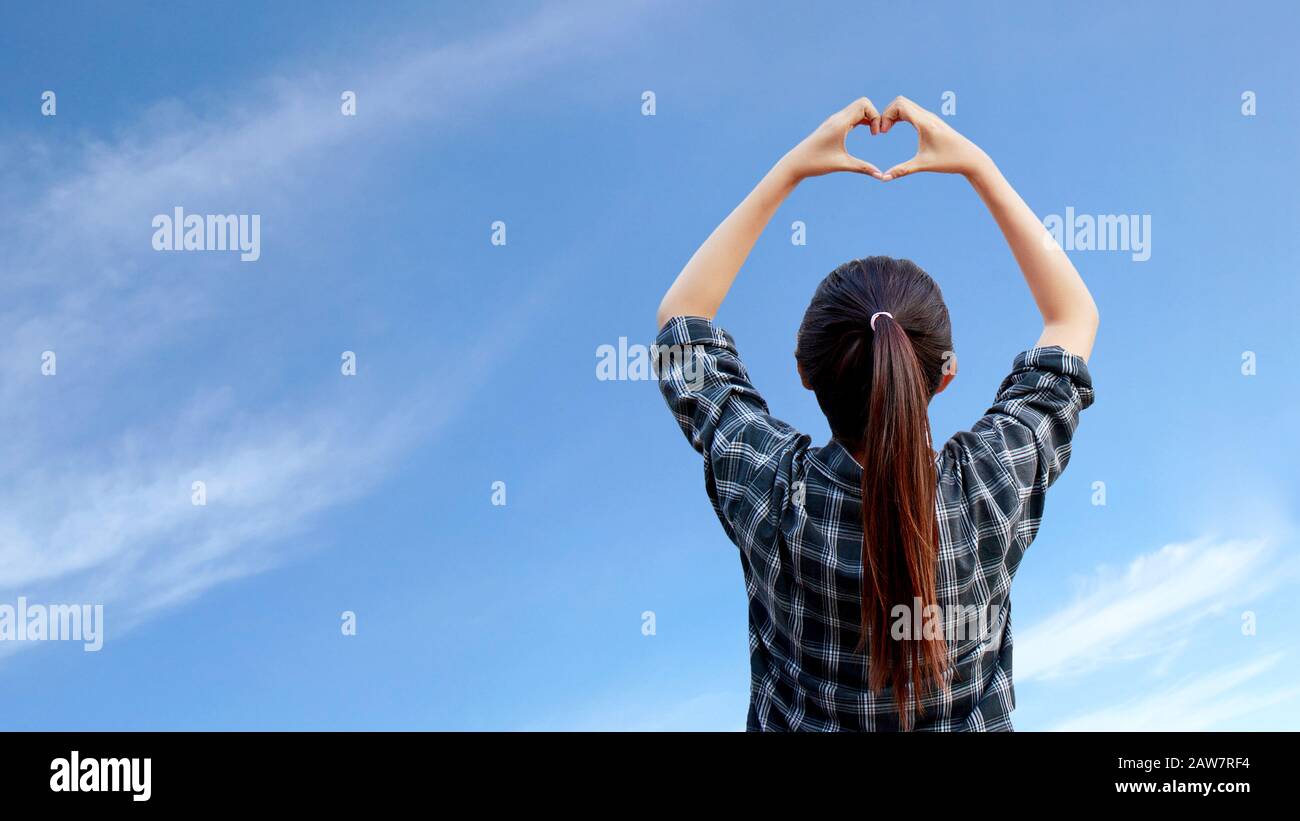 la mujer levanta las manos para dar forma al corazón en el aire con el cielo azul al fondo Foto de stock